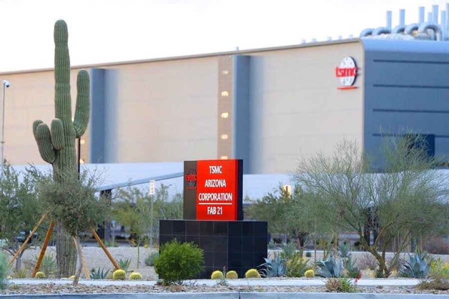 Industrial facility exterior with large beige and gray building featuring TSMC logo on side multiple tall green cacti in foreground red sign reading TSMC Arizona Corporation Fab 21 on black base surrounded by desert shrubs yellow flowers and chain-link fencing under clear sky