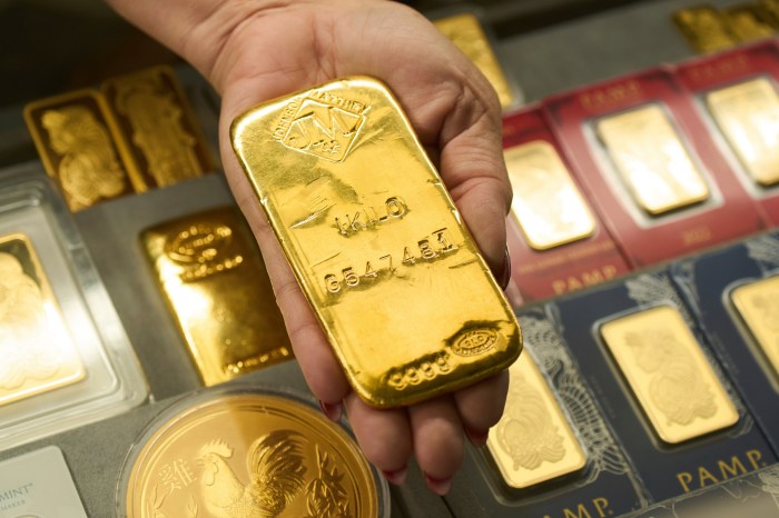 A hand holds a gold bar, with other gold bars and coins visible in the background.