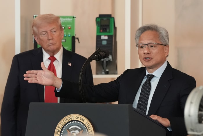 Nvidia CEO Jensen Huang speaking at a White House event, with President Trump in the background.