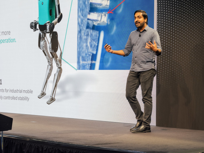 Pras Velagapudi speaking at the Humanoids Summit with a robot illustration on screen.