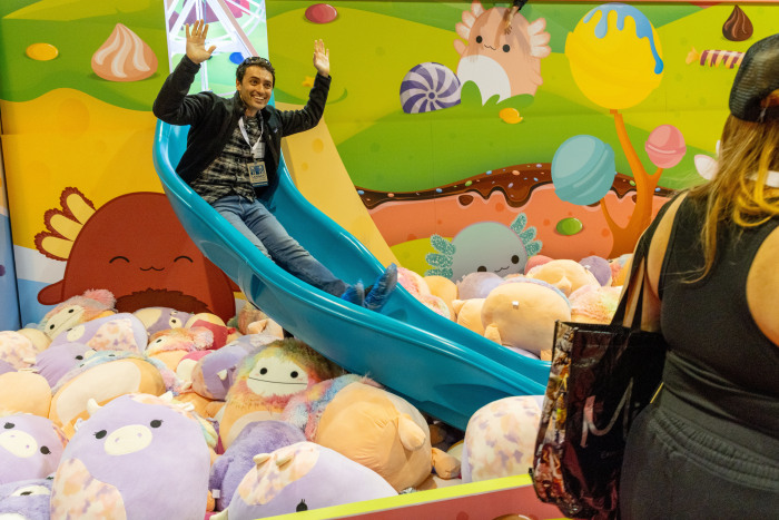A man slides down a blue slide into a pit of Squishmallow stuffed animals.
