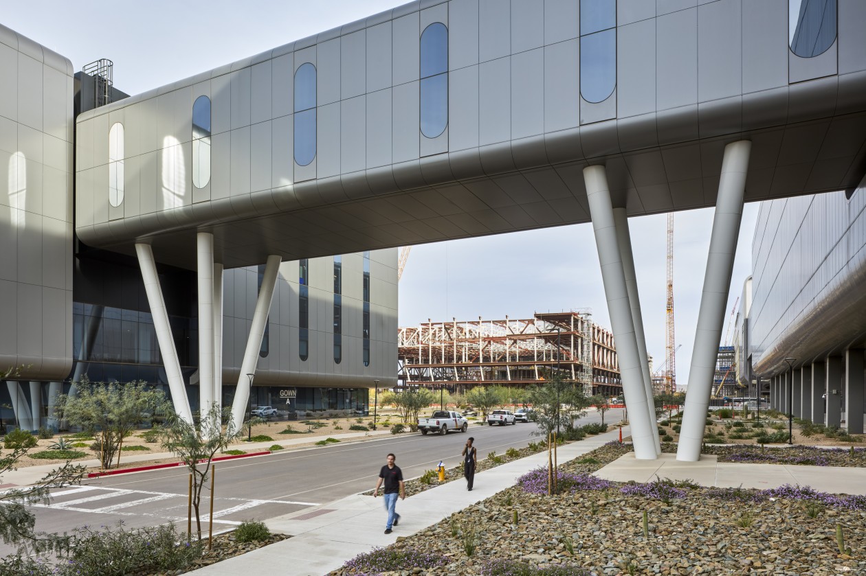 Two people walk along a sidewalk under a skybridge connecting two modern buildings in Phoenix.