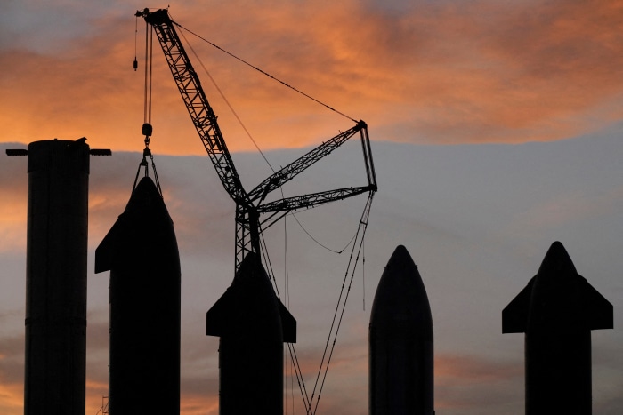 Silhouettes of SpaceX Starship prototypes and a crane against an orange and gray sky.