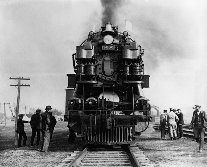A Union Pacific steam locomotive, number 9000, with smoke billowing from its smokestack, facing the viewer on train tracks as men stand on either side.