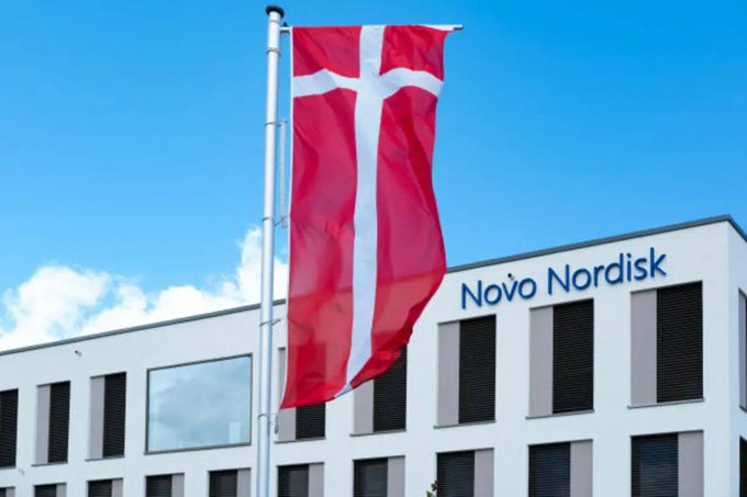A modern building with the name Novo Nordisk in blue text on its facade. A Danish flag with a red background and white cross is flying on a flagpole in front of the building. The sky is clear and blue with some white clouds.